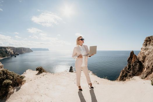 Successful business woman in yellow hat working on laptop by the sea. Pretty lady typing on computer at summer day outdoors. Freelance, travel and holidays concept.