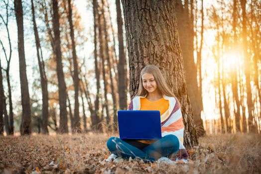 Autumn park with bright colorful foliage and teenage girl sitting under the tree with a laptop