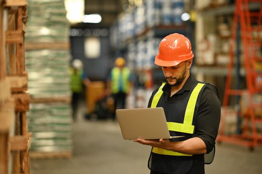 Warehouse manager wearing hardhats and reflective jackets using laptop for checking stock and order details in warehouse.