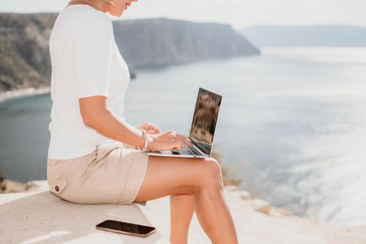 Successful business woman in yellow hat working on laptop by the sea. Pretty lady typing on computer at summer day outdoors. Freelance, travel and holidays concept.