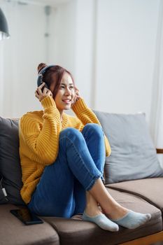 portrait of a young Asian woman woman using headphones holding smartphone while sitting on sofa.