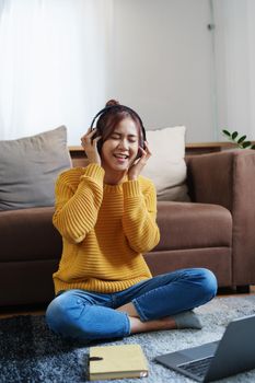 A portrait of a young Asian woman with a smiling face wearing a pair of headphones and using computer and listening to music while sitting on the sofa.