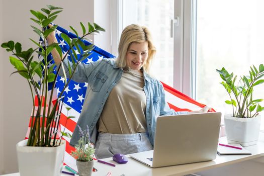 Happy woman employee sitting wrapped in USA flag, shouting for joy in office workplace, celebrating labor day or US Independence day. Indoor studio studio shot isolated on yellow background.