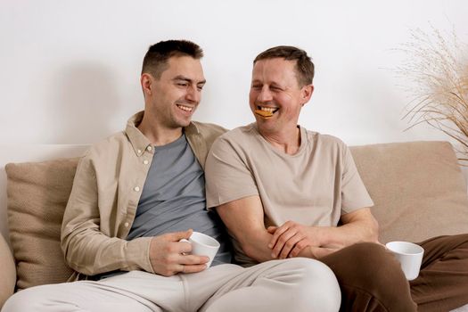 Happy gay couple with casual clothes spending time together at home, watching movie on the laptop and drinking tea. Two caucasian men relaxing. Homosexual relationships and alternative love