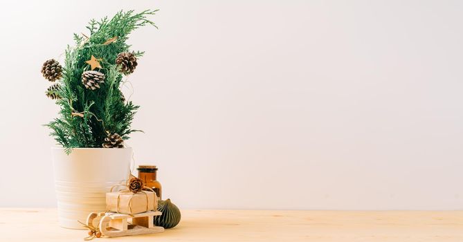 Creative natural miniature Christmas tree in pot on wooden table with gift boxes and white background. Xmas greeting card with copy space. Happy New Year