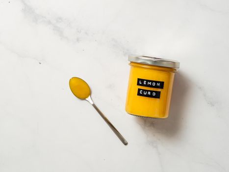 Delicious homemade lemon curd. Glass jar with lemon curd and full spoon on white marble background. Black sticky label with text Lemon Curd on jar. Top view or flat lay. Copy space