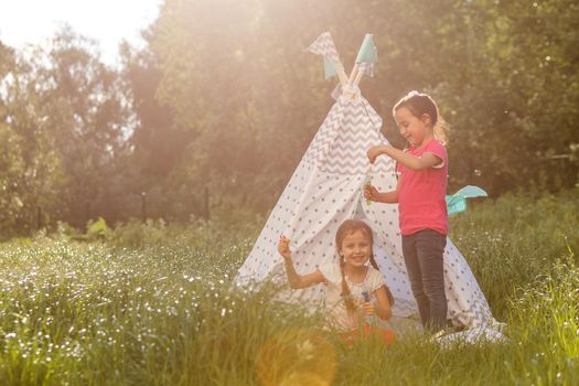 Two happy little kids are sitting on the grass and reading fairy tales near a tent in their home yard. Home camping vacation concept.