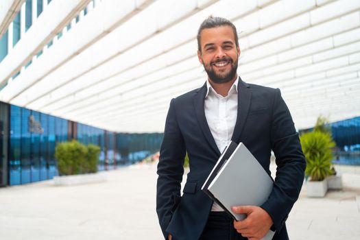 Successful businessman in suit with beard standing in front of office building confidently looking at camera and smile holding document. Hispanic male business person.