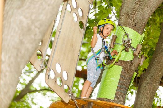 Portrait of little smiling girl in helmet and harness on trail in sky rope park in summer