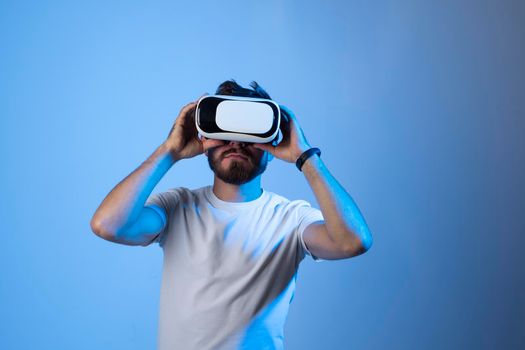 Bearded man in a white t-shirt wearing VR glasses and speaking with a friends in metaverse in a room with blue light. VR helmet for entertainment, learning and gaming