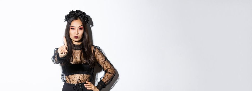 Serious-looking sexy asian woman in black gothic dress celebrating halloween, showing stop gesture, shaking finger to prohibit or refuse something, standing over white background.