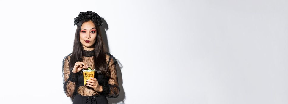 Confident smiling asian woman looking satisfied, holding sweets and wearing witch costume on halloween, standing over white background.