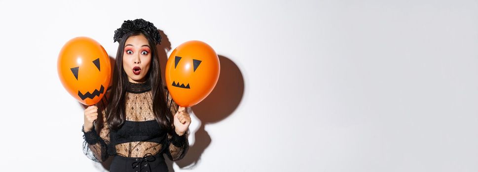 Image of surprised asian woman in witch costume celebrating halloween, holding balloons with scary faces, standing over white background.