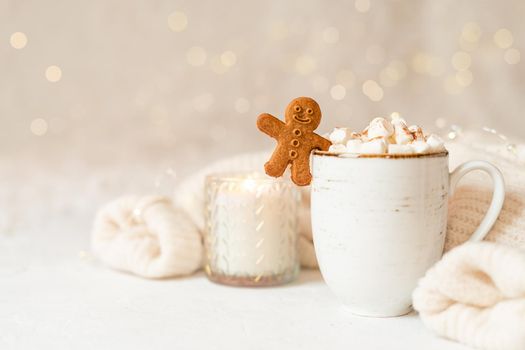 Gingerbread man cookie in cup of hot cocoa or coffee with marshmallow, candle, cinnamon and warm cozy sweater. Christmas lights background. Xmas holiday wooden star decorations.