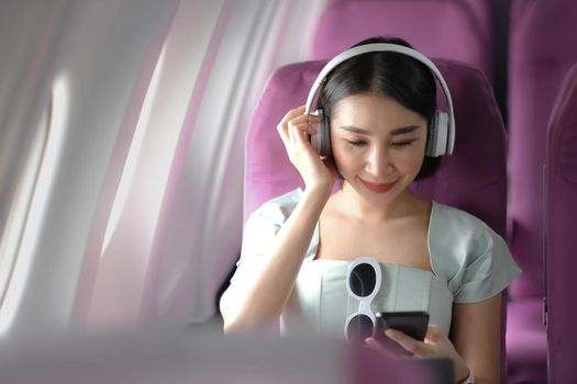 Travel tourism with modern technology and air flights concept, woman sitting in plane with modern smartphone and searching favourite music playlist in application for listening.