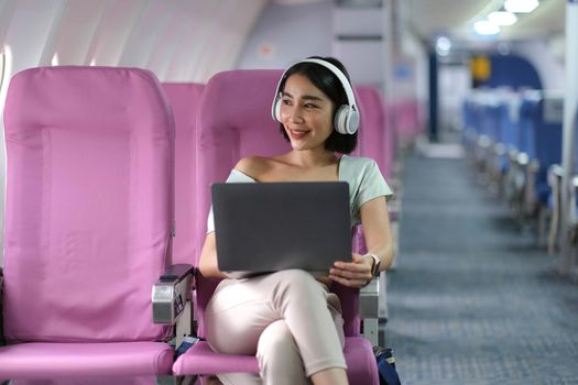 Asian young woman wearing headphone using laptop sitting near windows at first class on airplane during flight, Traveling and Business concept.