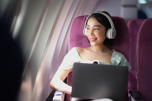 Asian young woman wearing headphone using laptop sitting near windows at first class on airplane during flight, Traveling and Business concept.