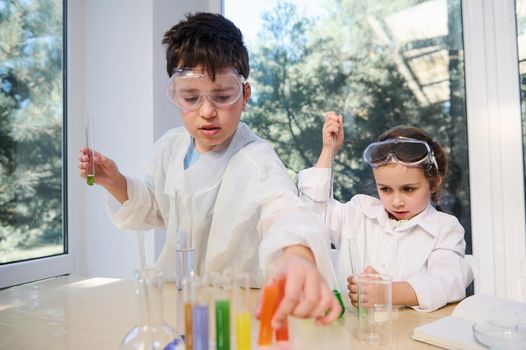 Cute kids, Caucasian boy and girl in protective eyewear and lab coats, learning Chemistry in chemical laboratory. Little children at lesson. Back to school. Elementary age students doing experiments