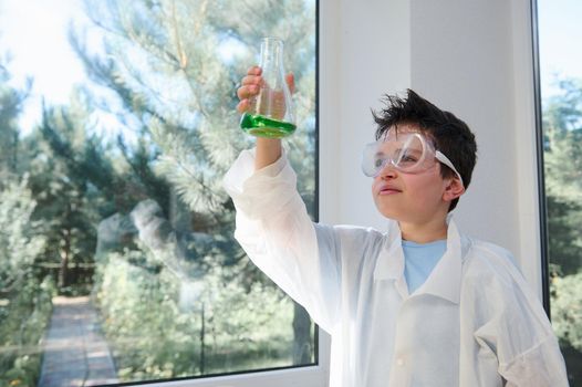 Adorable Caucasian dark-haired child, a smart schoolboy wearing safety goggles and lab coat, examining the chemical reaction going on in the flask. Learning Chemistry on new semester of a school year