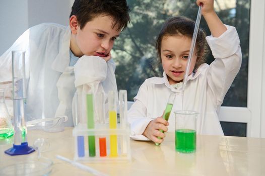 Smart elementary age students, adorable kids wearing safety goggles and white lab coat, watching a chemical reaction taking place in the test tube, while learning chemistry in the school laboratory