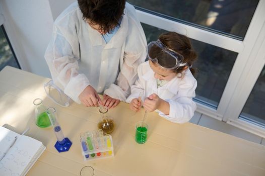 Top view of smart kids pupils, chemist scientist in white lab coats, holding labware with a taking place chemical reaction between chemicals and reagents, conducting experiments in chemistry classroom