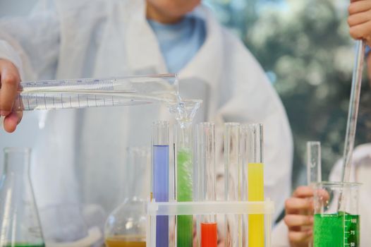 Selective focus on pouring water from measuring cylinder into test tubes with colorful chemical liquids, during chemistry lesson. Cropped blurred schoolboy conducting chemical experiments in classroom
