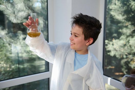 Happy mischievous smart schoolboy wearing lab coat, examining the chemical reaction going on in the flat bottomed flask. Learning Chemistry on new semester of academic year. Back to school. Education