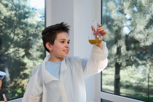 Smart child pupil, school boy chemist scientist in white lab coat, holding a flask with a taking place chemical reaction between chemicals and reagents, conducting experiments in chemistry classroom