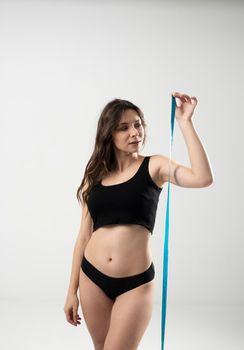 Confused brunette female with perfect slim body in a black underwear looking on a blue measure tape. Healthy nutrition and weight losing concept