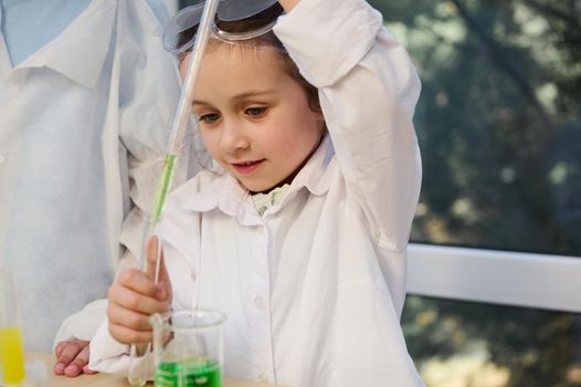 Smart Caucasian schoolgirl, elementary age student wearing white lab coat and goggles, conducts a chemical experiment in school Chemistry laboratory. Education, kids entertainment and development