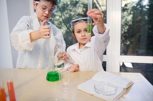 Beautiful Caucasian baby girl analyzing test tube with green chemicals and handsome smart child boy using a measuring pipette pipetting few reagents into a flat bottomed flask, during Chemistry lesson