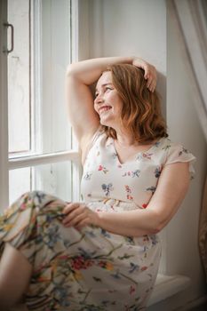 A middle-aged woman in a cream dress sits mysteriously and looks out the window on the windowsill. Green trees outside