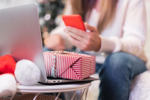 Close up woman with mobile phone, present gift box and laptop. Online shopping at Christmas holidays. Girl having a video chat at home and texting message. Merry Christmas and happy new year.