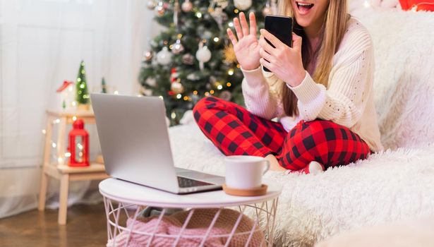 Happy smiling woman with gift box having a video call chat or taking selfie with her mobile phone at Christmas holidays. Online messaging with laptop at home. Happy new year and merry Christmas.