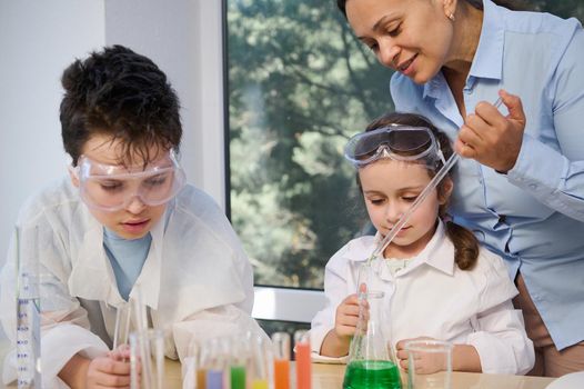 Charming African American young woman, experienced teacher, tutor explains a chemistry class to adorable smart children, conducting fascinated chemical experiments, in the school science laboratory