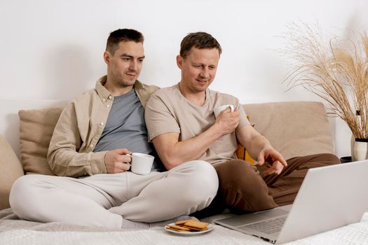 Happy gay couple with casual clothes spending time together at home, watching movie on the laptop and drinking tea. Two caucasian men relaxing. Homosexual relationships and alternative love