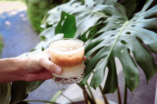 Hand hold iced latte coffee in a  tall glass. cold summer drink  with green nature background