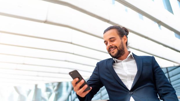 Businessman standing outdoor use smartphone looking device screen and smile. Handsome hispanic male business person in business suit holding smartphone in hand. Modern business person low angle view