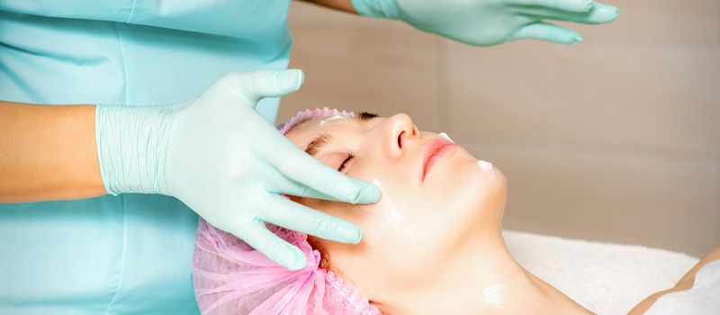 Cosmetologist with gloved hands applies a moisturizing mask with peeling cream on the female face. Facial cosmetology treatment. Procedures for facial care