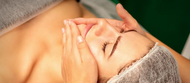 Beautiful caucasian young woman receiving a facial massage with closed eyes in spa salon, close up. Relaxing treatment concept