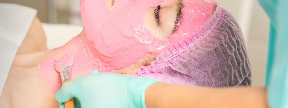 Hand of cosmetologist applying the pink alginic mask with the brush to the face of the young woman in a beauty salon