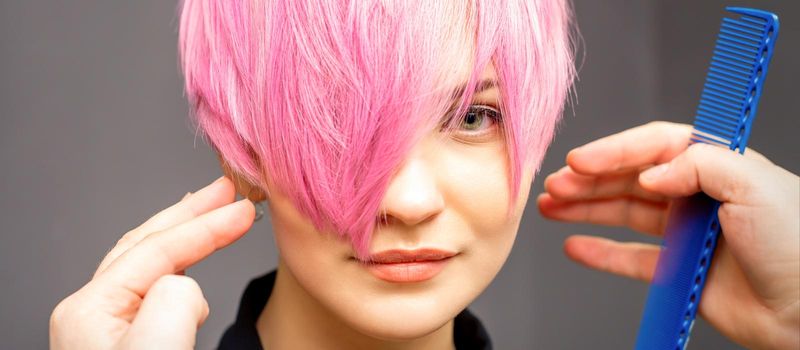 Hairdresser with hands and comb is checking out and fixing the short pink hairstyle of the young white woman in a hair salon