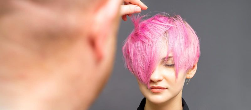 Hairdresser with hands is checking out and fixing the short pink hairstyle of the young white woman in a hair salon