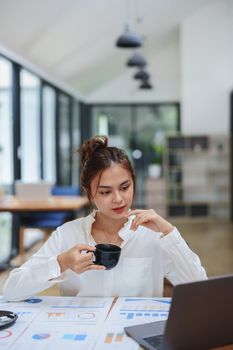 Portrait of a beautiful young woman working on a computer, documenting budget and drinking coffee during a review of the company's investment and earnings financial statements on her desk