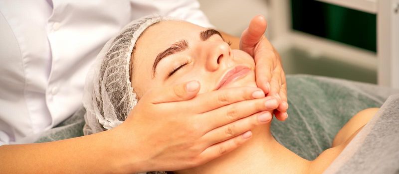 Beautiful caucasian young woman receiving a facial massage with closed eyes in spa salon, close up. Relaxing treatment concept