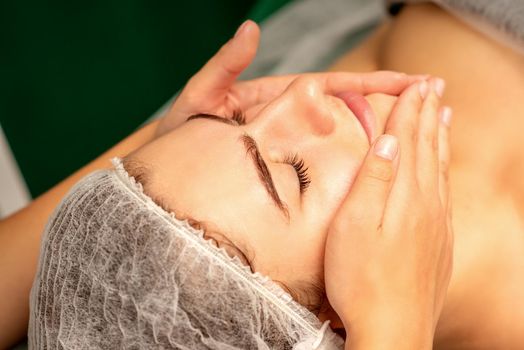 Beautiful caucasian young woman receiving a facial massage with closed eyes in spa salon, close up. Relaxing treatment concept