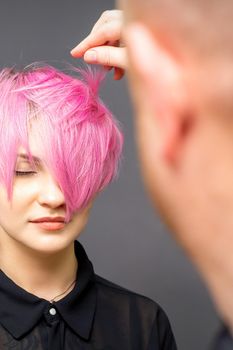 Hairdresser with hands is checking out and fixing the short pink hairstyle of the young white woman in a hair salon