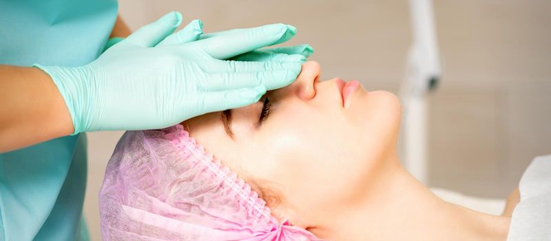 Cosmetologist with gloved hands applies a moisturizing mask with peeling cream on the female face. Facial cosmetology treatment. Procedures for facial care