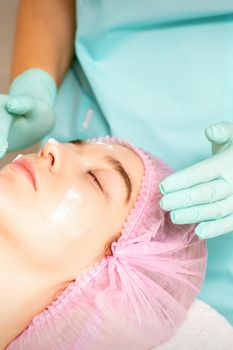 Cosmetologist with gloved hands applies a moisturizing mask with peeling cream on the female face. Facial cosmetology treatment. Procedures for facial care