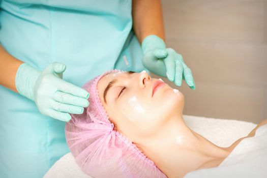 Cosmetologist with gloved hands applies a moisturizing mask with peeling cream on the female face. Facial cosmetology treatment. Procedures for facial care